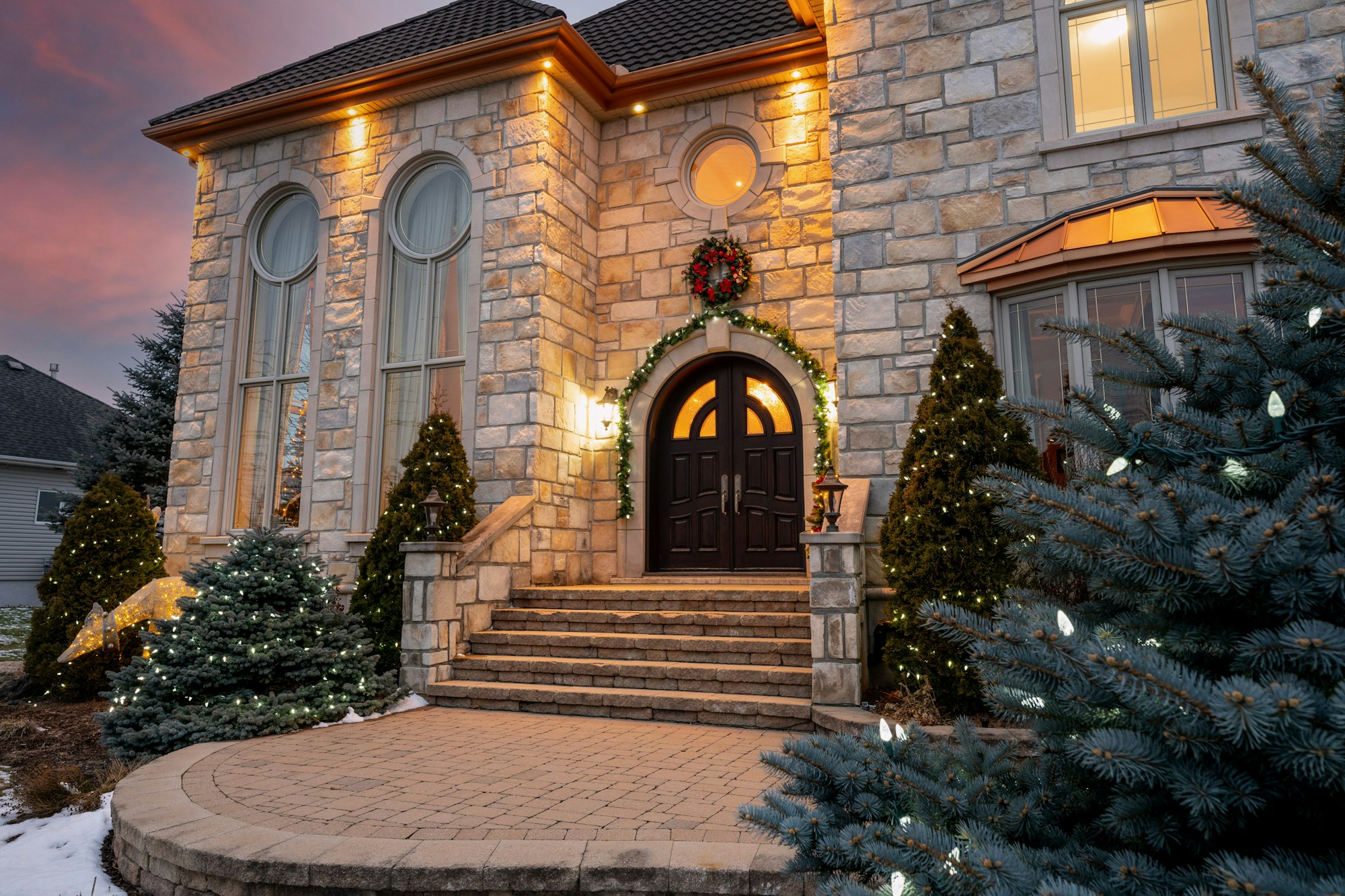 A luxury stone home entrance decorated with holiday lights and greenery at dusk